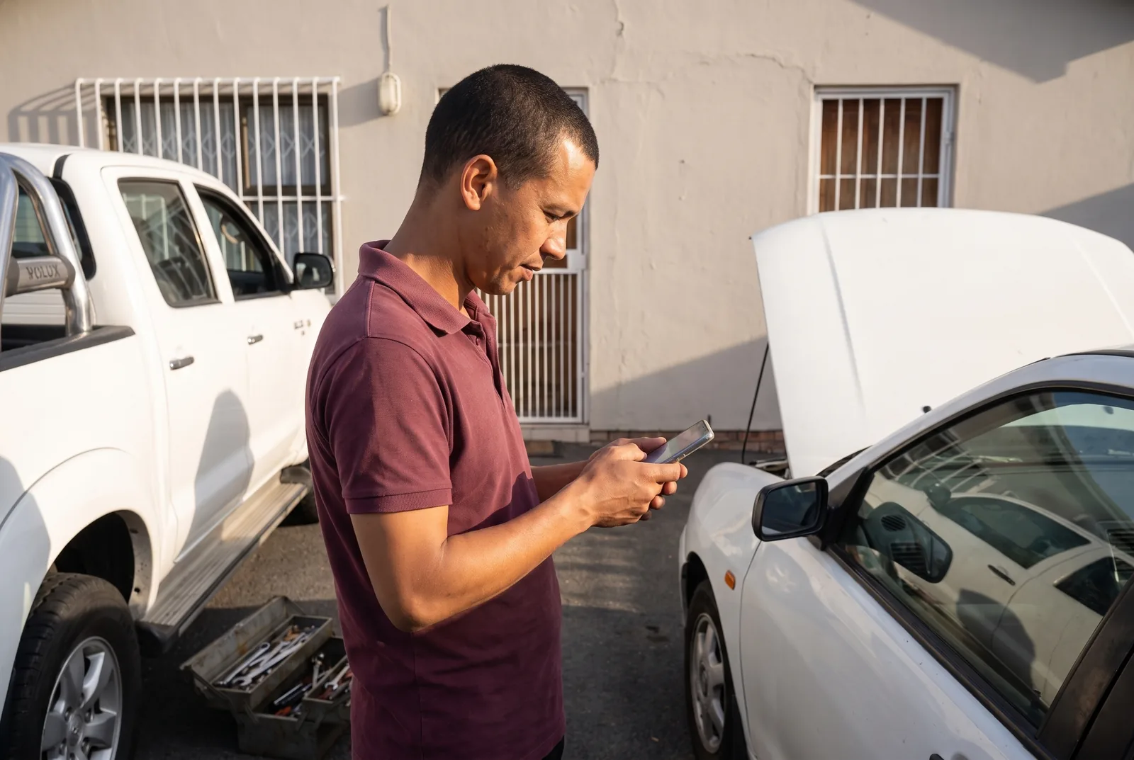 A mobile mechanic checking a customer's details on his phone in a suburban driveway, his bakkie and open toolbox behind him and the customer's car with its bonnet up beside him.
